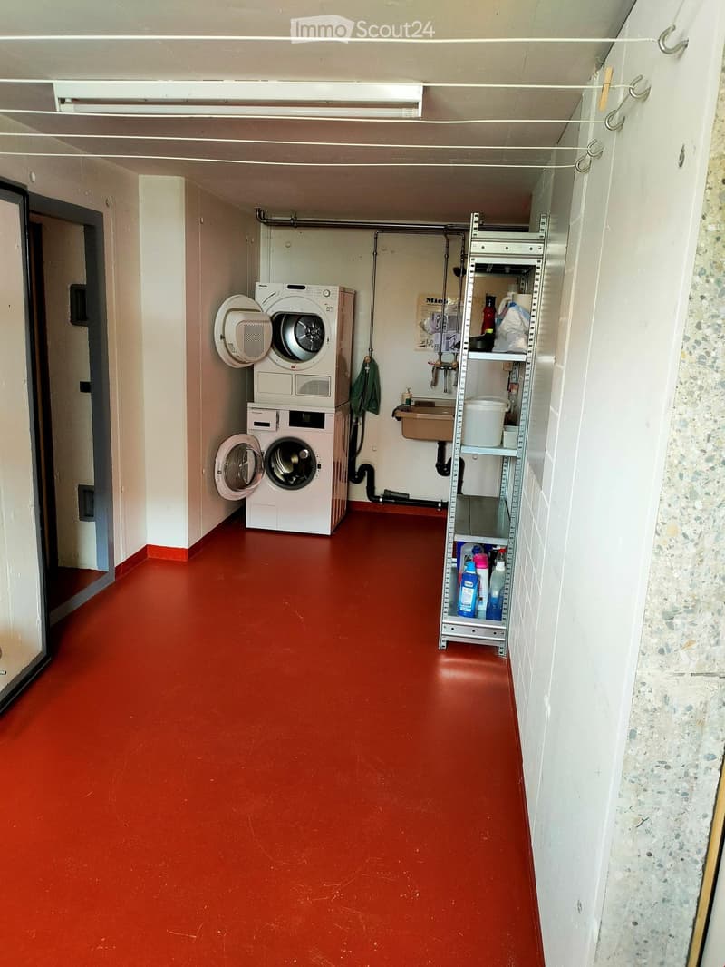 A narrow hallway with a red floor, white walls, and a washing machine and dryer unit on the left side. A metal shelf with cleaning supplies is on the right side.