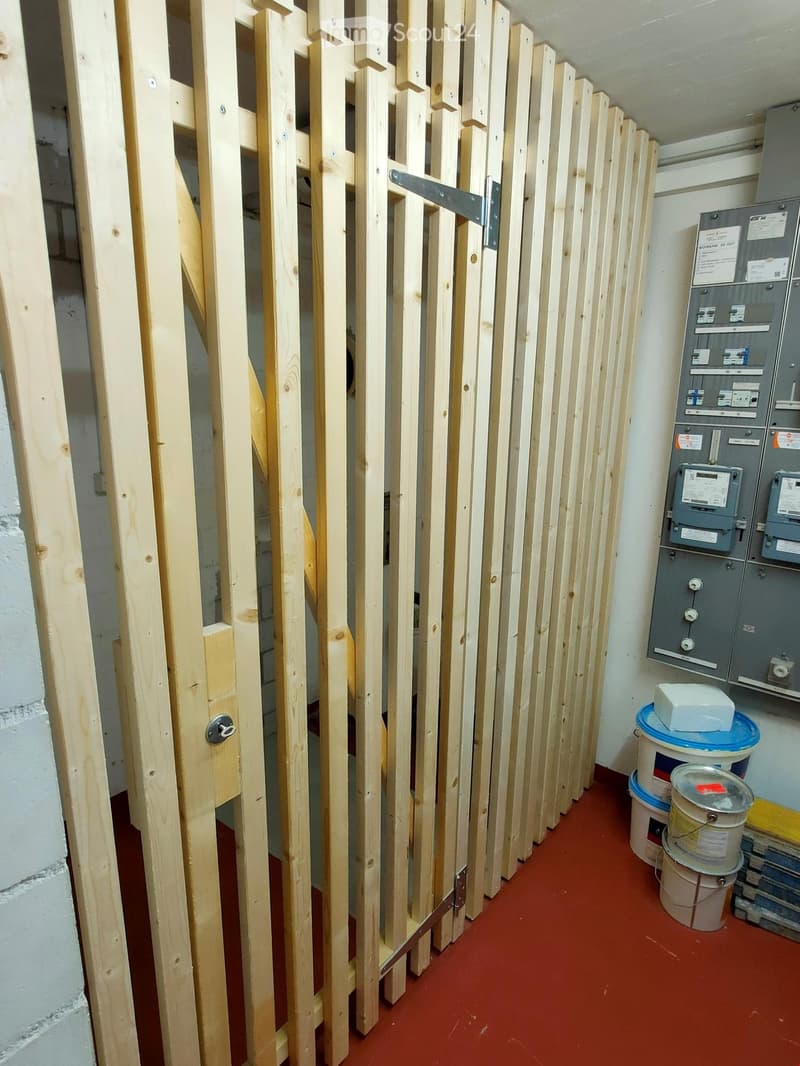 A narrow hallway with a red floor, white walls, and a wooden barrier on one side. Electrical panels and storage containers are visible on the right side.