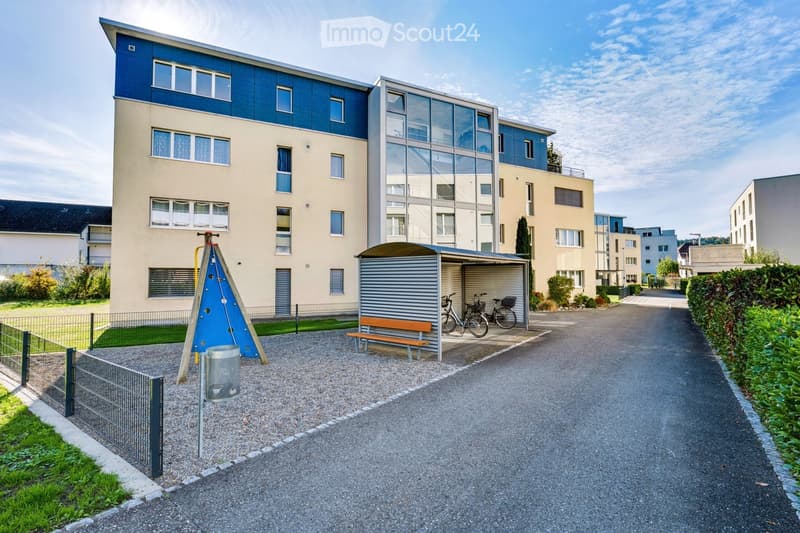 Modern multi-story residential building with a mix of beige and blue facades, large windows, and a covered bike parking area with bicycles and a bench.