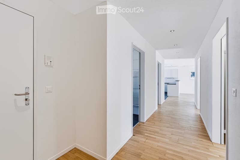 Bright, minimalist hallway with light wooden flooring, white walls, and recessed ceiling lighting. Features a white door on the left and an open doorway leading to a kitchen area on the right.