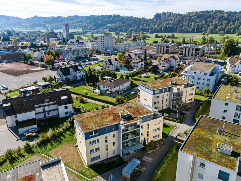 Aerial view of a residential area with multiple buildings, green spaces, and a surrounding landscape of hills and fields.