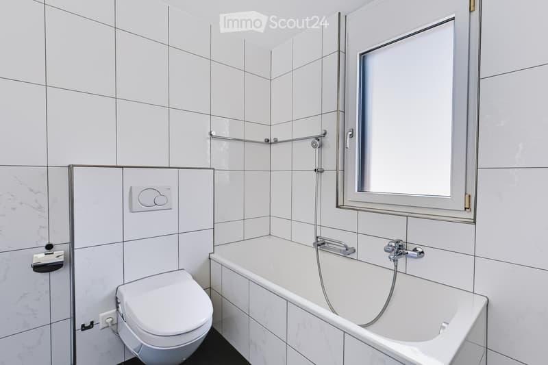 Modern bathroom with white tiles, a bathtub, toilet, showerhead, and a large mirror above the sink area.