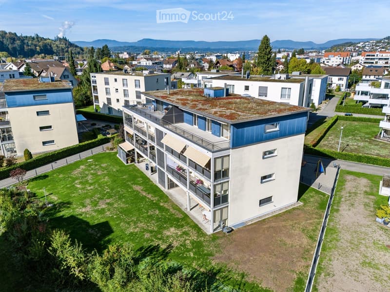 Modern multi-story residential building with balconies, green roof, and surrounding greenery in a suburban neighborhood.