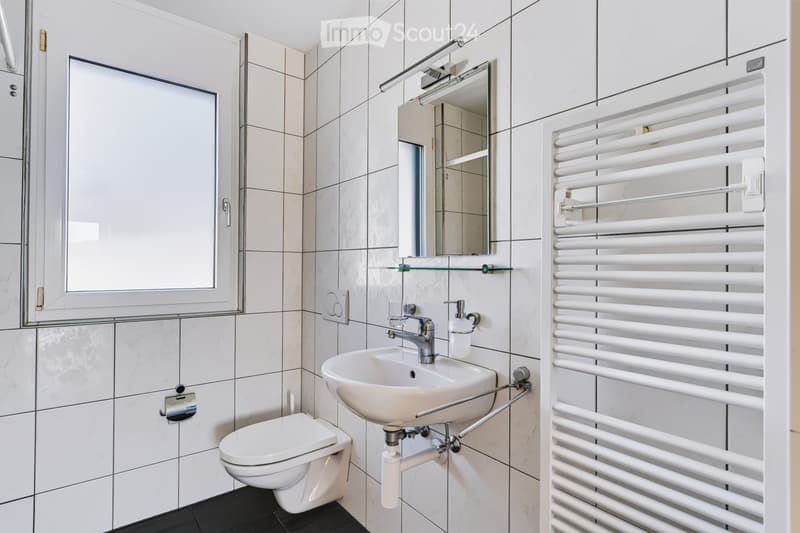 Modern bathroom with white tiles, a sink with a chrome faucet, a toilet, a large mirror with built-in lighting, and a white radiator.