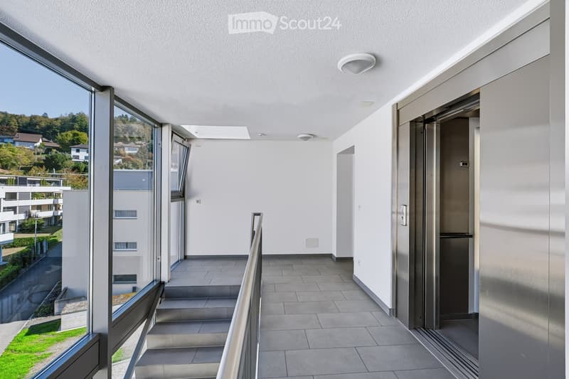 Modern hallway with large windows, elevator, and staircase leading to a view of the surrounding area.