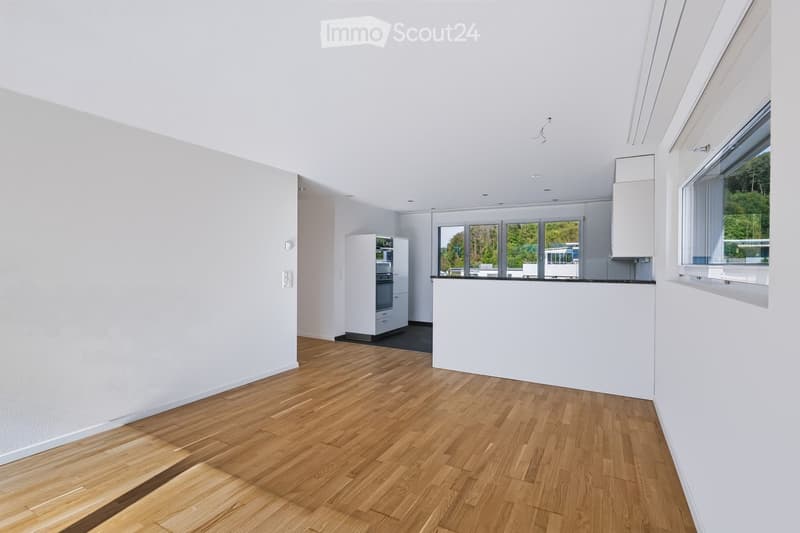 Modern kitchen with light-colored wooden flooring, white walls, and built-in cabinets. Features a refrigerator, oven, and large windows with a view of greenery outside.