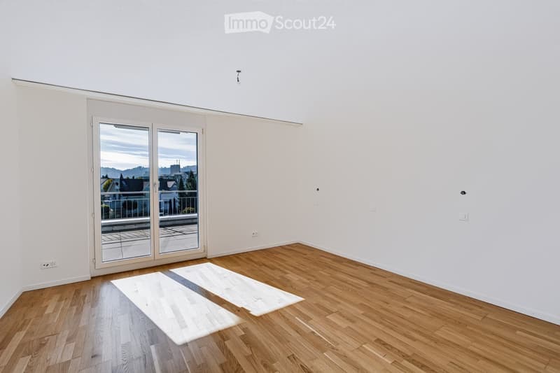 Empty room with wooden flooring, white walls, and large sliding glass doors leading to a balcony with a view of the city and surrounding landscape.