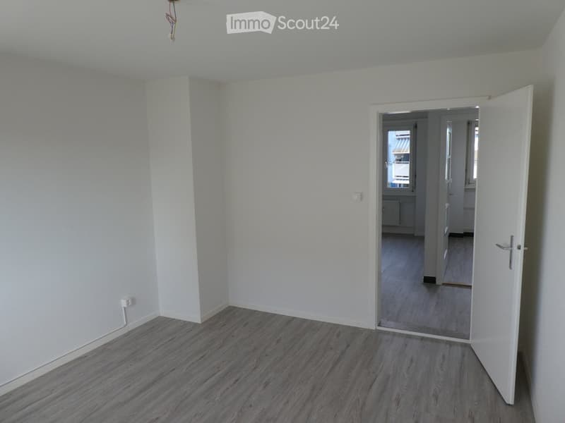 Empty room with light grey walls and wooden flooring, featuring an open doorway leading to another room.