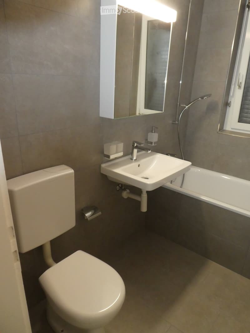 Modern bathroom with a toilet, sink, bathtub, and mirror. Features a sleek design with grey tiles and a window for natural light.