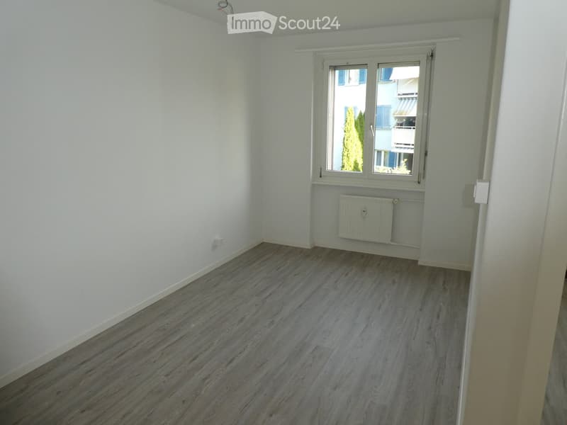Empty room with light wooden flooring, white walls, and a window with blinds. A radiator is visible below the window.