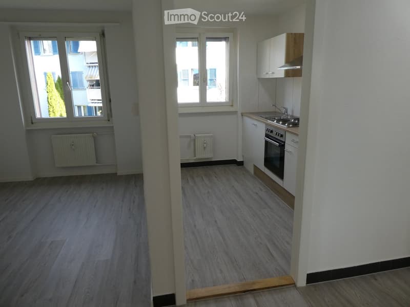 Empty room with light wooden flooring, white walls, and a small kitchen area featuring white cabinets, a sink, and an oven. Windows provide natural light.