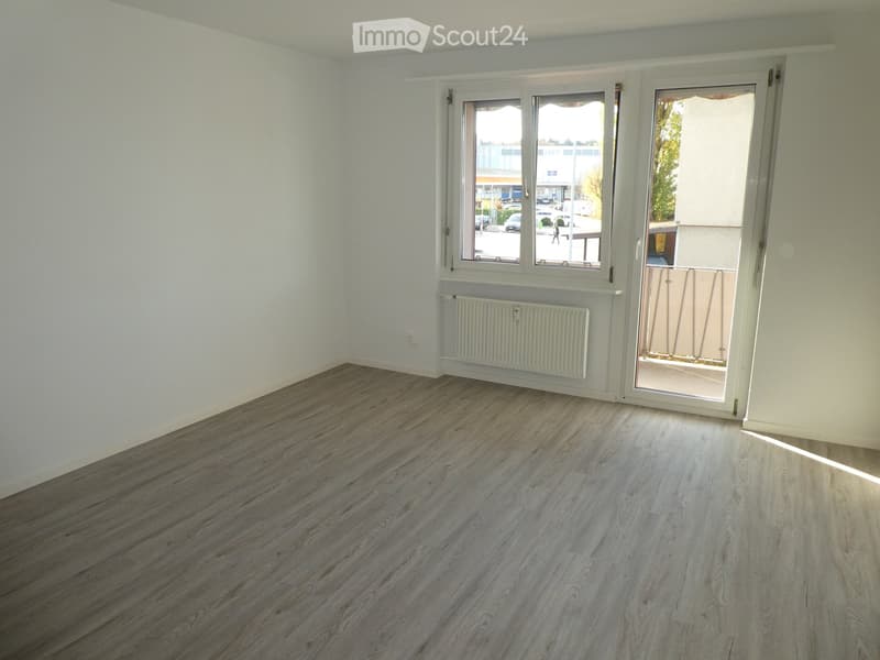 Empty room with light-colored walls, wooden flooring, a window, a radiator, and a sliding glass door leading to a balcony.