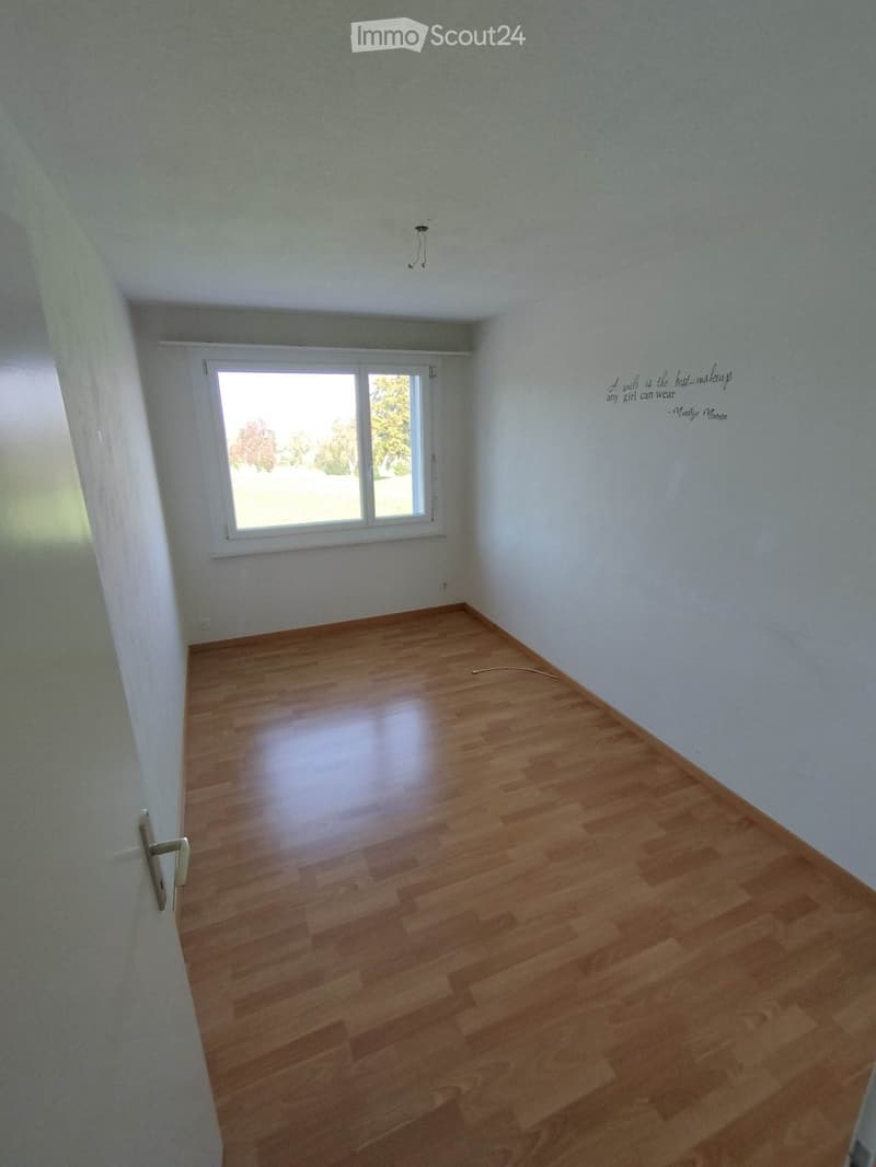 Empty room with wooden flooring, white walls, and a large window letting in natural light. The room has a quote written on the wall and a visible electrical outlet.