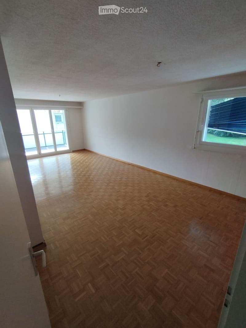 Empty room with wooden parquet flooring, white walls, and large windows, including a sliding glass door leading to a balcony.