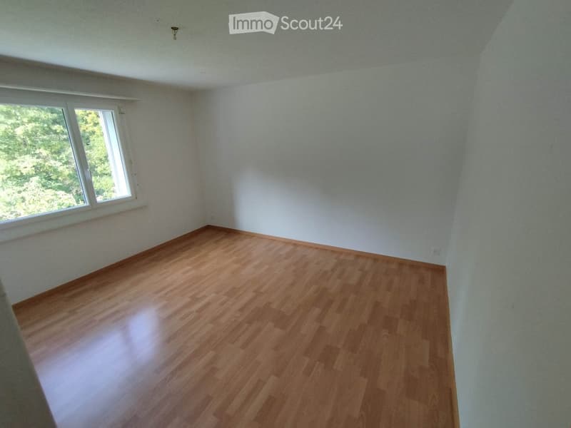Empty room with wooden flooring and white walls, featuring a large window with a view of greenery outside.