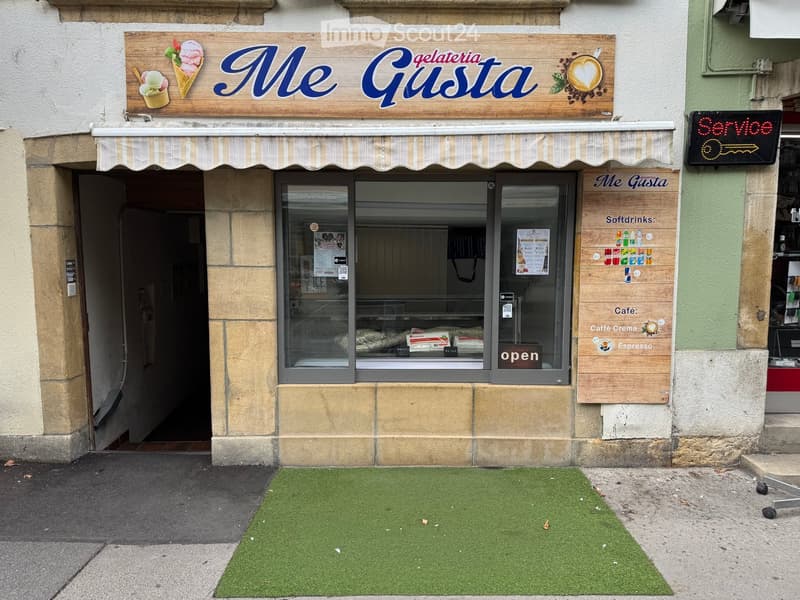Facade of a gelateria named "Me Gusta" with a striped awning, glass display window, and menu board listing soft drinks and coffee options.