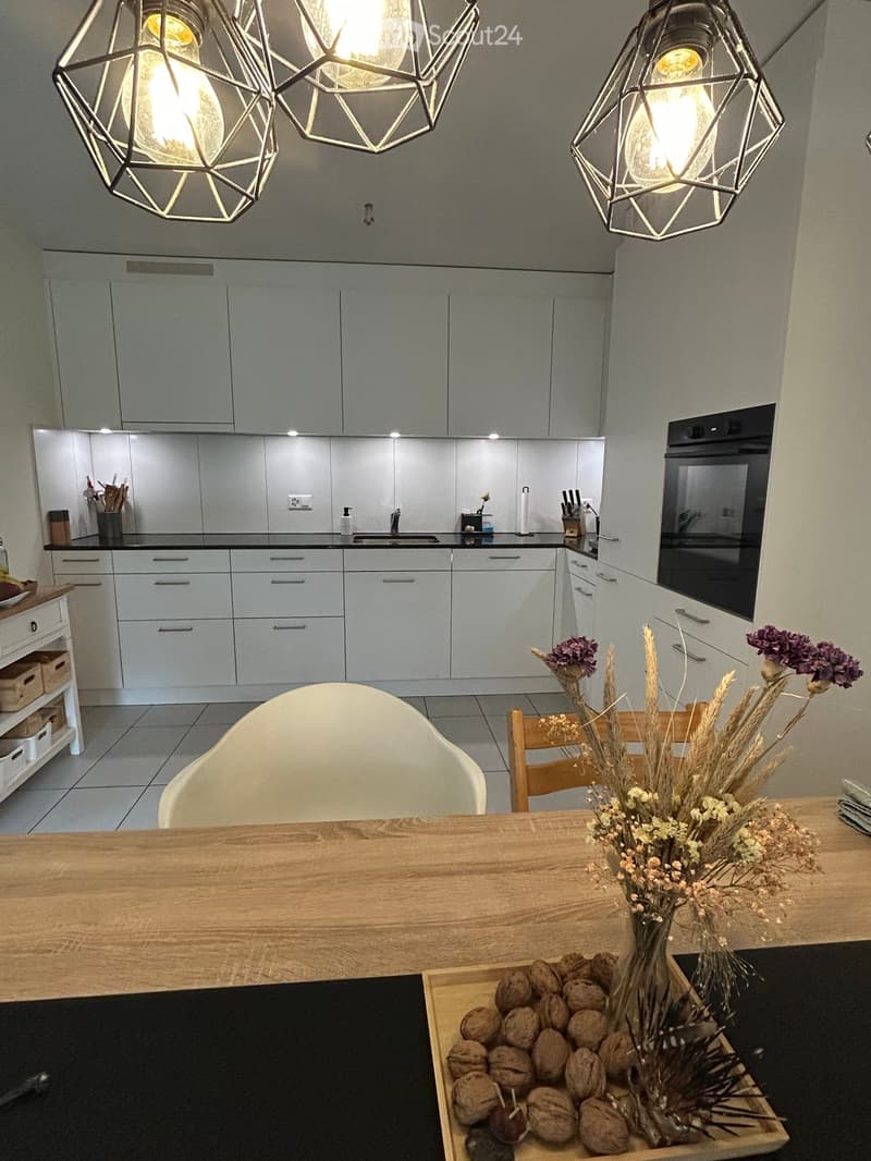 Modern kitchen with grey cabinets, a wooden dining table, white chairs, and a vase with dried flowers on the table. The kitchen features a sink, oven, and various utensils on the counter.