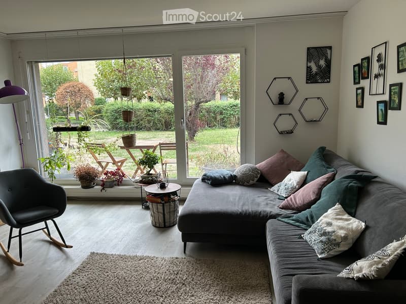 Modern living room with large windows overlooking a garden, featuring a grey sectional sofa with colorful pillows, a rocking chair, a coffee table, a floor lamp, and several potted plants on the windowsill.