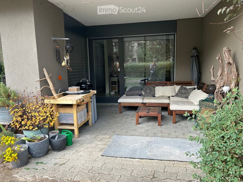 A well-furnished outdoor patio with a sofa, wooden table, potted plants, and a wooden wind chime. The space features a stone-paved floor and a view of the surrounding greenery.