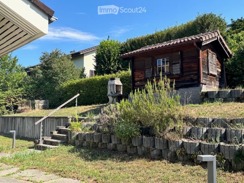 A small wooden house with a tiled roof and stone steps leading up to it, surrounded by greenery and a stone retaining wall.