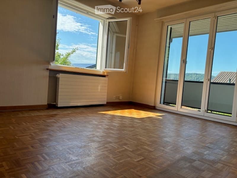 Empty room with wooden flooring, large windows, radiator, and sliding glass doors leading to a balcony with a scenic view.