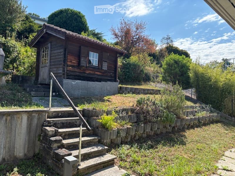 A small wooden house with a tiled roof, situated on a raised concrete platform with stone steps leading up to it. The house has a single window and a door, surrounded by greenery and shrubs.