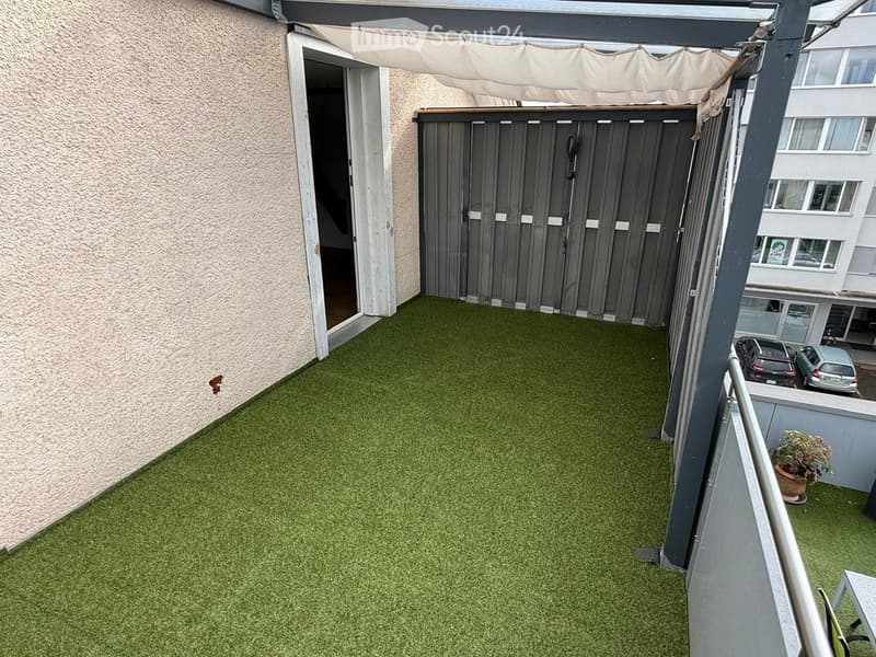 Balcony with artificial grass flooring, covered by a retractable awning, adjacent to a building entrance.