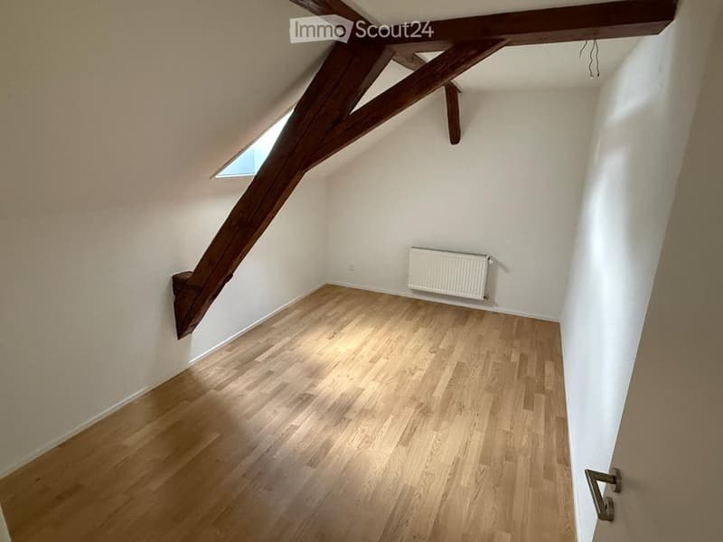 Empty attic room with wooden floor, white walls, exposed wooden beams, and a small skylight window.