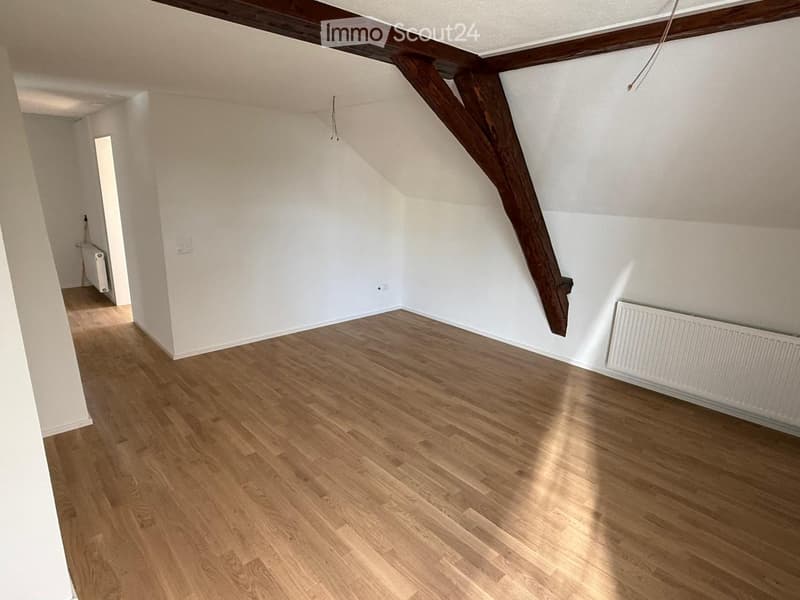 Empty attic room with wooden floors, white walls, exposed wooden beams, and radiators on both sides.