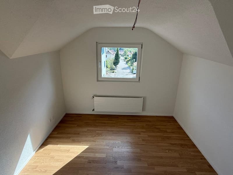 Empty attic room with wooden flooring, white walls, a single window showing a street view, and a radiator below the window.