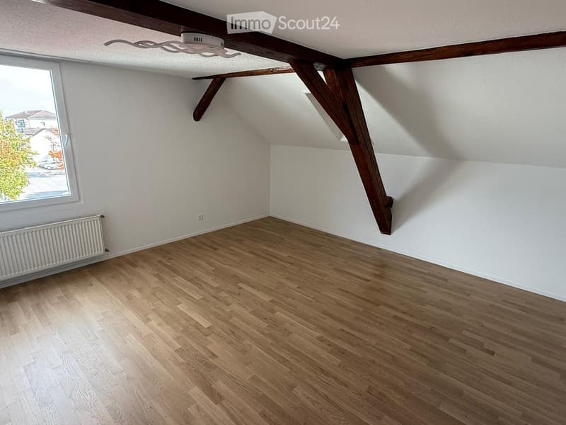 Empty attic room with wooden beams, wooden flooring, white walls, and a window with a view of the outside.