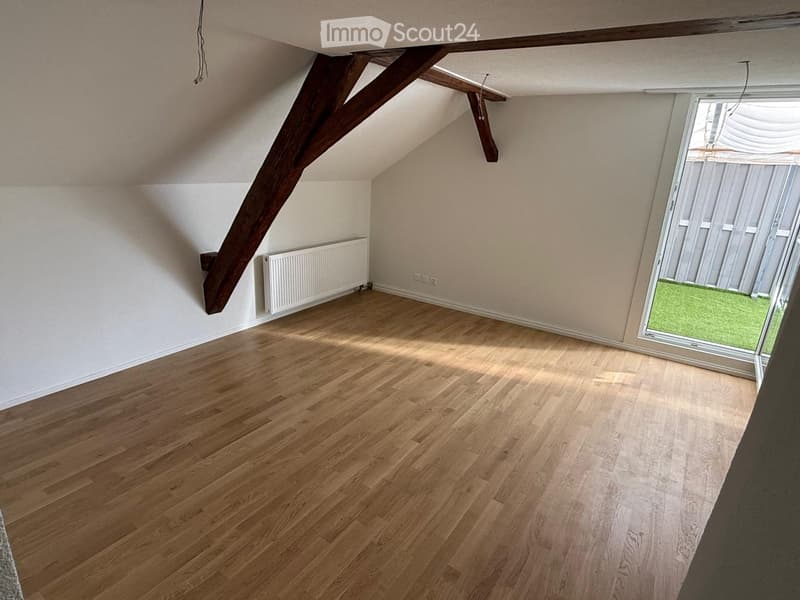Empty attic room with wooden flooring, exposed wooden beams, white walls, radiator, and a window opening to a green outdoor area.