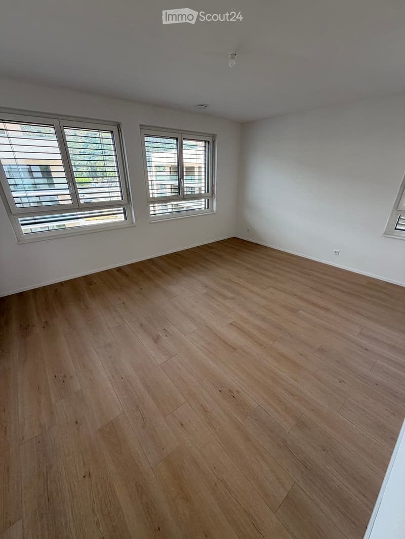 Empty room with light wooden flooring, white walls, and large windows with blinds.
