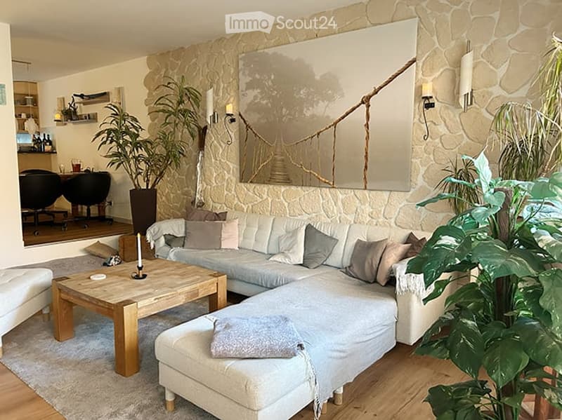 Modern living room with a large sectional sofa, coffee table, ottoman, and a large mirror on a stone accent wall. The space includes a wooden floor, a potted plant, and a small dining area visible in the background.