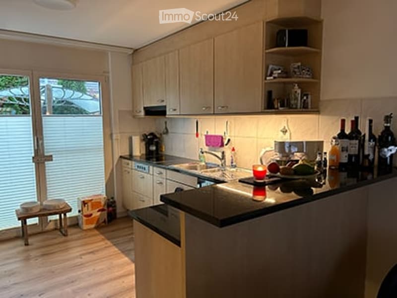 Modern kitchen with light wood cabinets, stainless steel appliances, and a black countertop. A small stool is near the window, and several bottles and a candle are placed on the counter.