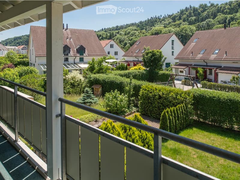 Balcony overlooking a well-maintained garden and neighboring houses with red-tiled roofs, surrounded by lush greenery and trees.