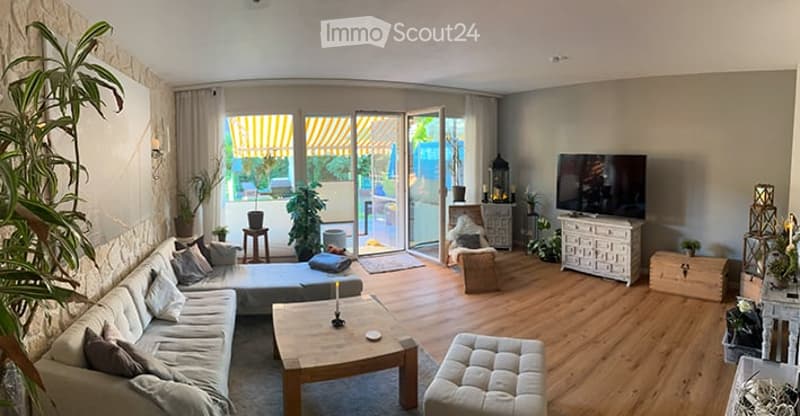 Bright living room with large sliding glass doors leading to a balcony, wooden flooring, a grey sectional sofa, coffee table, ottoman, TV, and decorative plants.