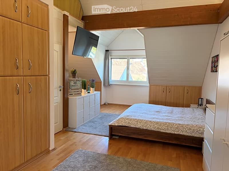 Cozy bedroom with wooden floors, slanted ceiling, large window, bed with patterned bedding, nightstand, TV mounted on wall, small cabinet with plants, and two area rugs.