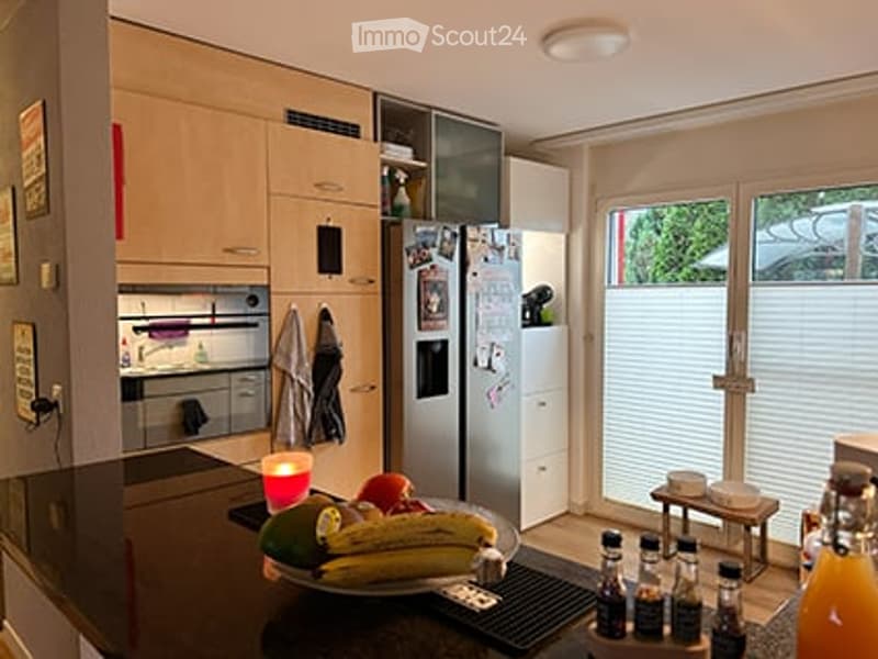 Modern kitchen with light wood cabinets, stainless steel appliances, and a black countertop. A bowl of fruit and several bottles are placed on the counter, along with a small stool near the window.