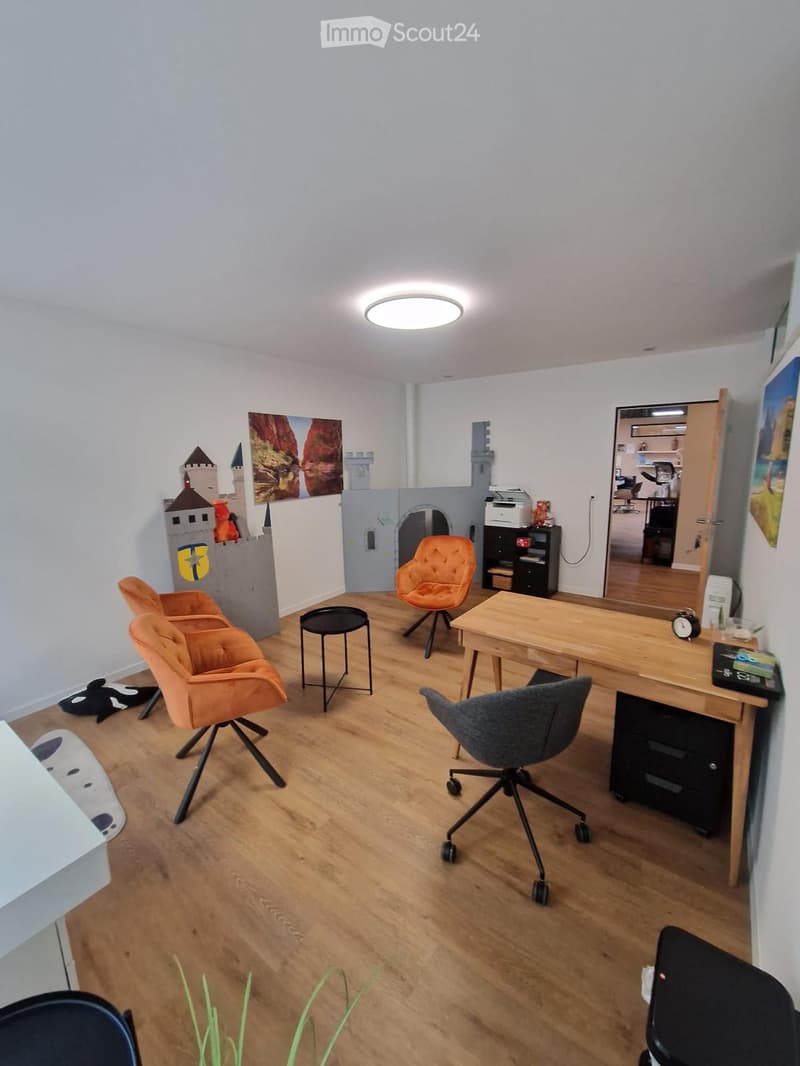 A well-lit room with wooden flooring, featuring a desk with a chair, a small table with a stool, and several colorful chairs. A toy castle and artwork are visible on the walls.