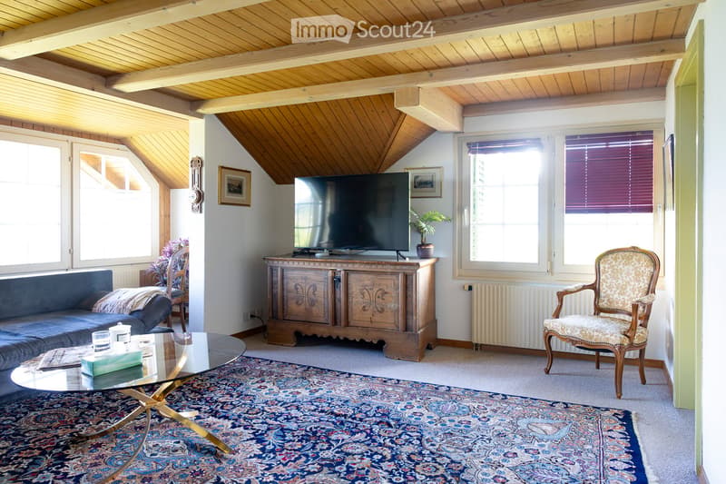 Living room with exposed wooden beams, large windows, a dark blue sofa, a glass coffee table, a television on a wooden stand, a patterned armchair, and a decorative rug.