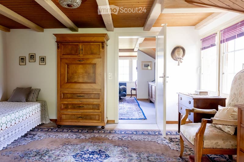 Cozy attic bedroom with exposed wooden beams, a daybed, a wardrobe, a chair, and a small table with a lamp.