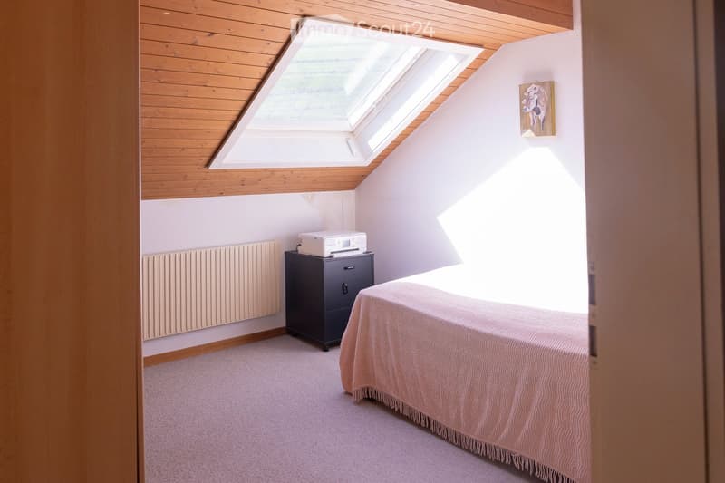 Bright attic bedroom with wooden slanted ceilings, skylight window, bed with pink bedding, nightstand with printer, radiator, and a picture frame on the wall.