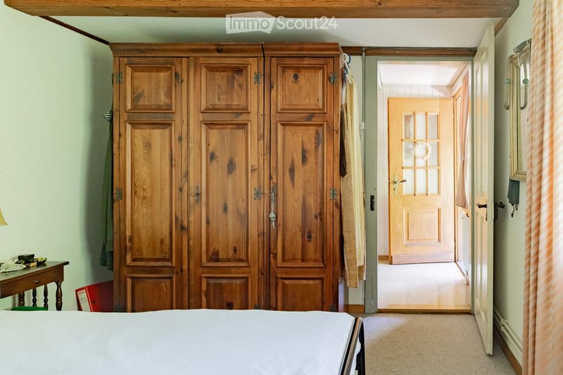 Cozy bedroom with a large wooden wardrobe, a neatly made bed, a small wooden table with a lamp, and a mirror on the wall. The room features light-colored walls and carpeted flooring, with a doorway leading to another room.