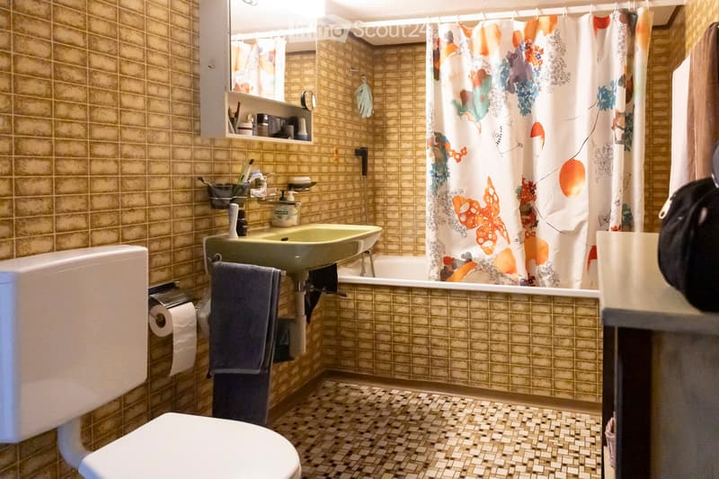 Bathroom with beige tiled walls and floor, featuring a toilet, sink, bathtub, and shower curtain with a floral design. A towel hangs on a rack, and a shelf holds toiletries.