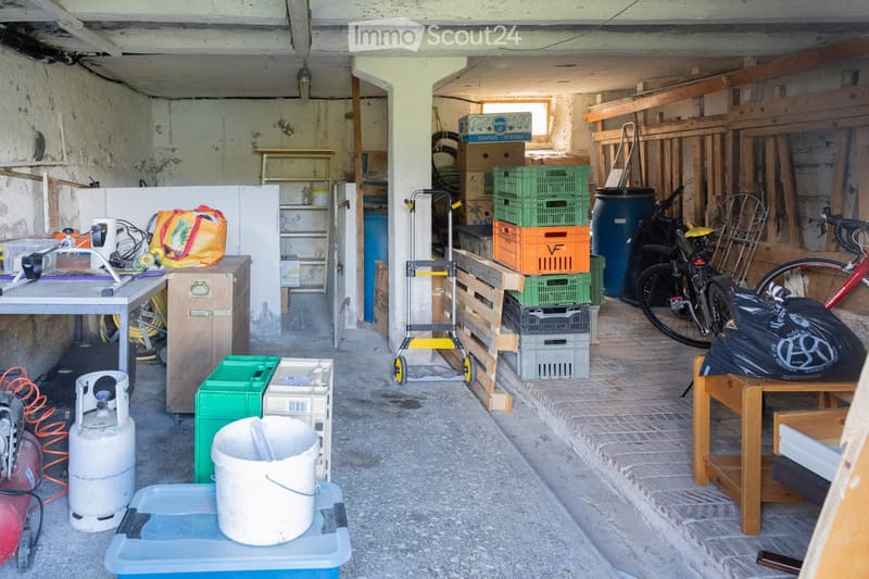 The image shows a cluttered garage or storage area with various items including a table, chair, bicycle, gas cylinder, handcart, crates, and a stool.