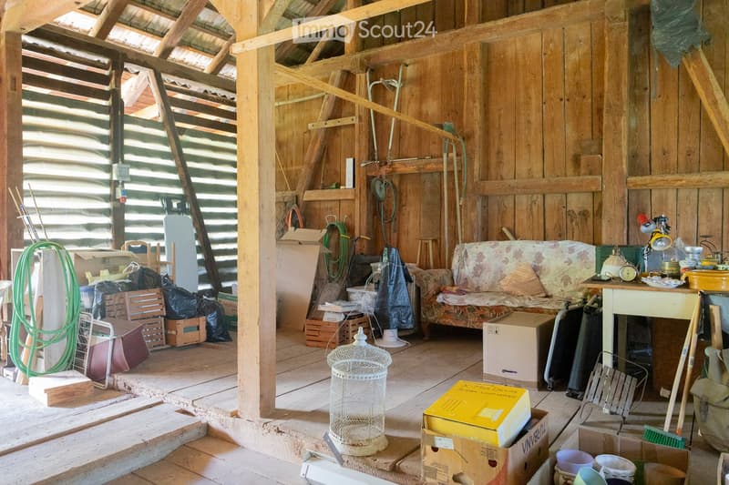 A rustic wooden room with exposed beams and a high ceiling, featuring a patterned sofa, a small table with various items, a birdcage, and several stools scattered around.