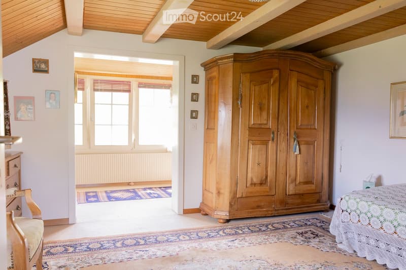 Cozy bedroom with wooden wardrobe, bed with lace cover, chair, table, and picture frames on the walls.