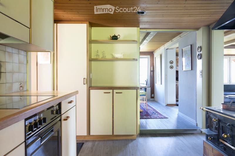 The image shows a cozy, well-lit kitchen area with light green cabinets, wooden countertops, and a modern oven. A small dining area with a table, chairs, and a rug is visible in the background, along with decorative items like a teapot and picture frames