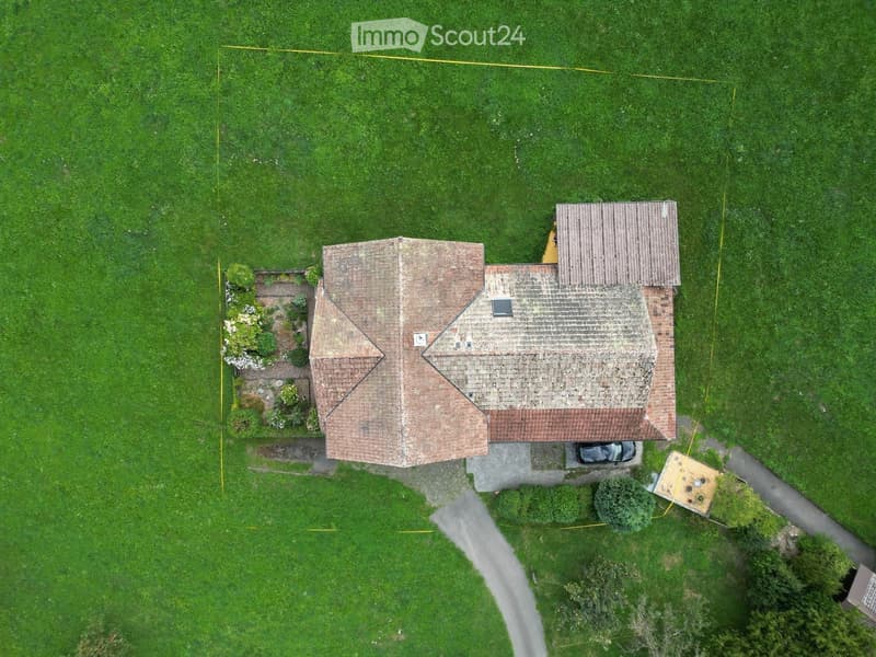 Aerial view of a two-story house with a tiled roof, surrounded by a well-maintained lawn and garden area. A car is parked in the driveway, and there is a small outdoor patio or deck to the right.